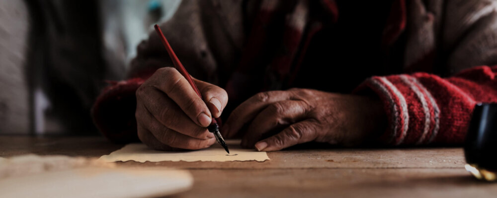 Old man with dirty hands writing a letter using a nib pen and ink or pigment from a pot in a low angle view across a wooden table.